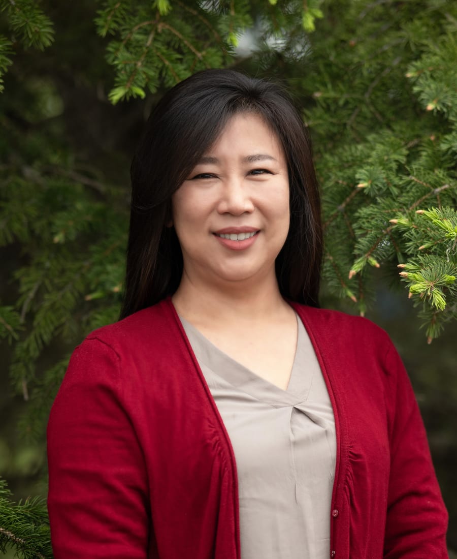 Eun Kyeong (Judy) Cho, smiling, in a red cardigan in front of a spruce tree